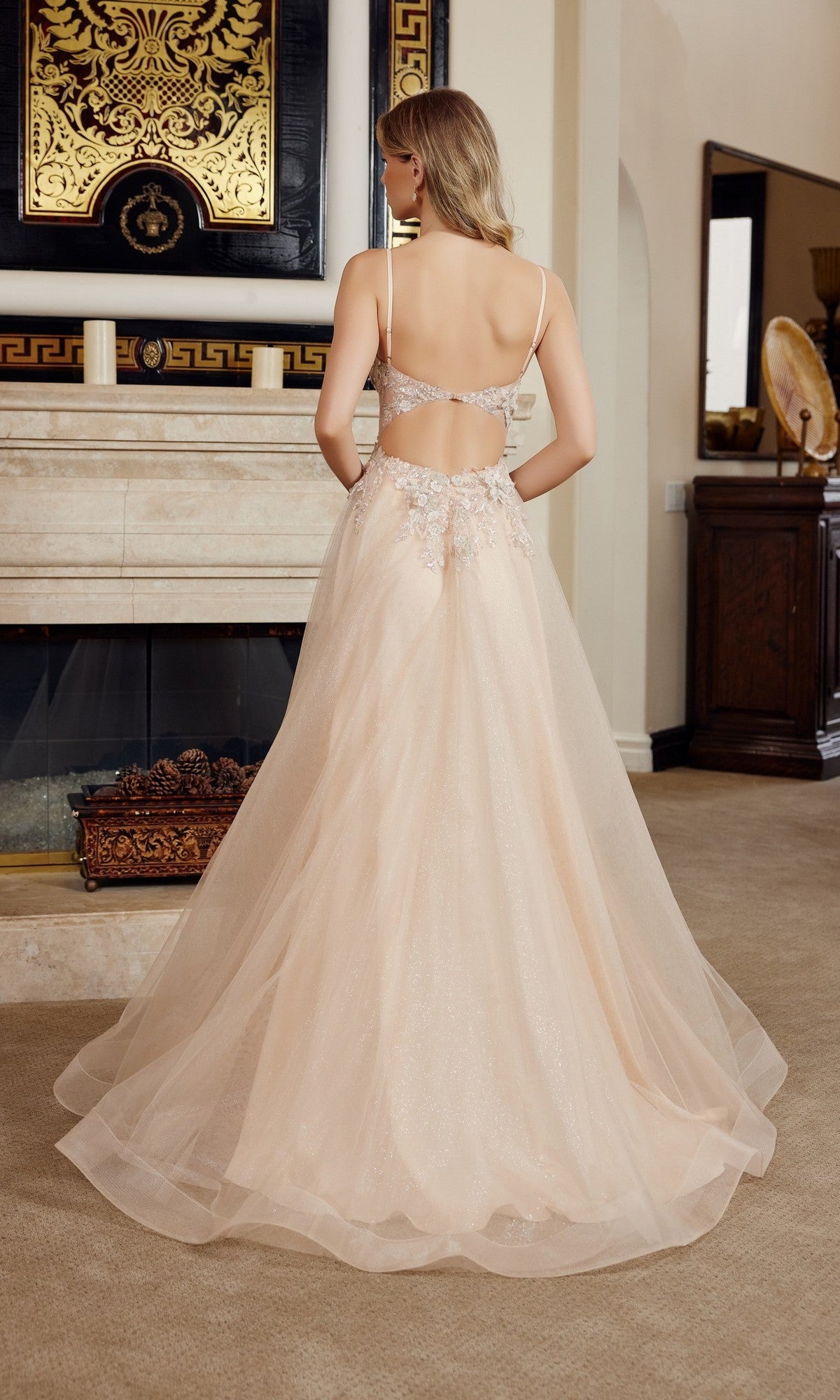 A woman wearing the Nox Anabel G1787, a light peach, floor-length sleeveless prom dress with an A-line silhouette and 3D floral appliques, stands facing a fireplace in a decorated living room.